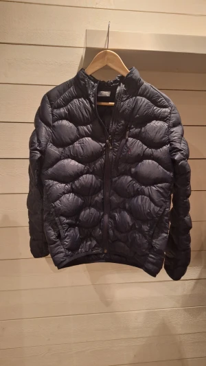 Svart dunjacka Peak Performance 160 - Säljer en svart dunjacka från Peak Performance i storlek 160. Jackan har quiltad design, hög krage och tvåvägsdragkedja framtill. Liten broderad logga på bröstet och två fickor med dragkedja. Perfekt för kyliga dagar och riktigt snygg passform.