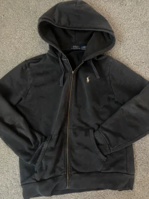 Svart hoodie från Polo Ralph Lauren - Svart hoodie från Polo Ralph Lauren med dragkedja framtill, huva med snörning och klassisk logga broderad på bröstet. Mjuk bomullskänsla och kängurufickor. Perfekt för chill dagar eller när du vill ha en clean streetstyle.