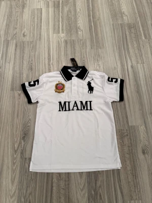 Vit Polo Ralph Lauren Miami piké - Vit pikétröja från Polo Ralph Lauren med svart krage och svarta detaljer. Stor broderad logga med polospelare, Miami-text och emblem på bröstet. Siffran 5 på ärmen och ryggen. Klassisk passform och tillverkad i bomull. Perfekt för dig som gillar sportig och stilren look.