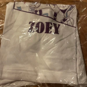 Vit croppad KPop Demon Hunters t-shirt ZOEY - Cool vit croppad t-shirt med lila anime-tryck och texten 'HUNTRX ZOEY' framtill. Kortärmad modell med rund hals och figurnära passform. Perfekt för KPop Demon Hunters fans! 