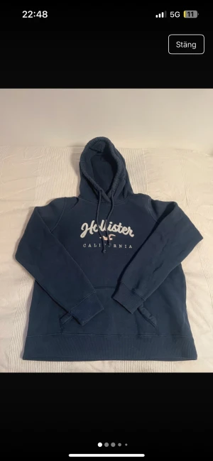 Marinblå Hollister hoodie med broderi - Snygg marinblå hoodie från Hollister med broderad vit logga på bröstet. Tröjan har huva med snörning, känguruficka och är tillverkad i mjuk bomullsmix med fleece på insidan. Perfekt för chill dagar och enkel att matcha med jeans.