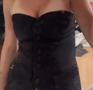 Svart korsett med knappar - Säljer en svart korsett med snygg bandeauform och stora knappar framtill. Toppen är axelbandslös och har en figurnära passform som framhäver midjan. Perfekt för dig som vill ha en stilren och trendig look.