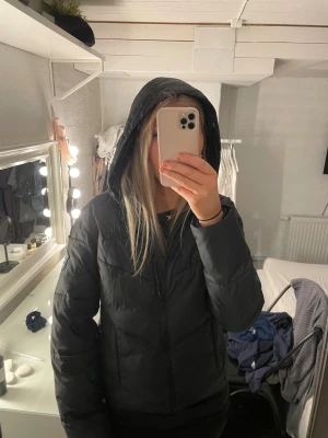 Svart pufferjacka med huva från Svea - Svart pufferjacka från Svea med broderad logga på bröstet och patch på ärmen. Jackan har huva, dragkedja framtill och två sidofickor. Materialet är syntet och jackan har quiltad design som ger en schysst look. Perfekt för kalla dagar. Nypriset är ca 1000kr.