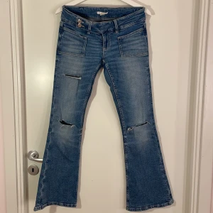 Blå bootcut jeans från Gina Tricot 14+ S - Bootcut jeans från Gina Tricot 14+ i blå tvätt med slitna detaljer och hål vid knäna. Modellen har låg midja, klassiska fickor fram och bak samt dragkedja och knapp. Jeansen är tillverkade i denim och har en snygg, avslappnad vibe. Hålen har spruckit lite.