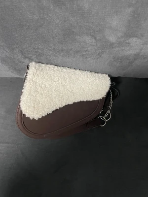 Brun axelväska med pälsdetalj - Säljer en snygg brun axelväska med mjuk vit pälsdetalj på locket. Väskan har en cool form, bred axelrem och silverfärgade ringar och kedjor som ger extra edge. Perfekt för dig som vill sticka ut med en unik accessoar. Materialet är skinn och pälsimitation.