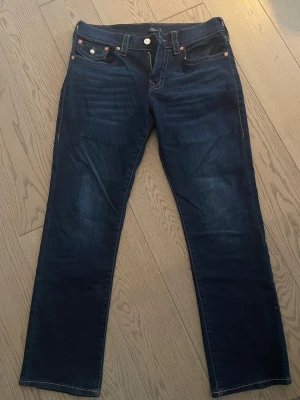 True Religion Ricky W33 - Här har vi de perfekta vardagsjeansen med snygga detaljer! Ricky jeansen från True Religion har en rak och avslappnad passform och är tillverkade av slitstark denim känd för sin hållbarhet. Designade med hästskodetaljerade fickor i äkta True Religion-stil.  Endast provade, i nyskick.  Myntficka Relaxed fit Dragkedja framtill Raka ben Bomullsblandning Denim Stretch 5 fickor Mid waist 98% Bomull, 2% Elastan