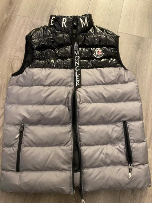 Moncler svart och grå pufferväst ( M storlek) - Snygg pufferväst från Moncler i svart och grått med glansig finish upptill och broderad logga på bröstet. Västen har hög krage med broderade bokstäver, två dragkedjefickor och stor Moncler-text längs dragkedjan. Perfekt för lager-på-lager och streetstyle. A-B+