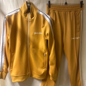 Palm Angels senapsgula tracksuit - Palm Angels mjukisbyxor i senapsgult med vita och svarta sidoremsor. Byxorna har elastisk midja, raka ben och logotyptryck på vänster lår. Materialet är mjukt och stretchigt, perfekt för chill dagar. Snygg streetwear-vibe med sportiga detaljer. 