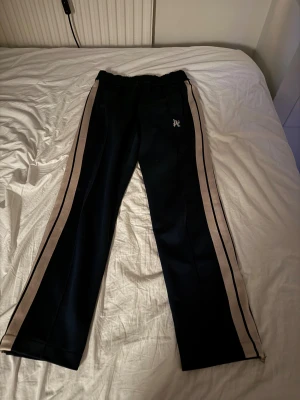 Palm Angels sweatpants med monogram - Palm Angels sweatpants i mörkblått med broderat monogram på framsidan. Byxorna har raka ben, beige sidoremsor och dragkedja vid benslut. Elastisk midja för skön passform. Liten defekt på vänster knä, men syns inte på långt avstånd. Priset går att diskutera!