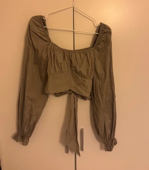 Beige omlottopp med puffärmar - Snygg beige omlottopp med långa puffärmar och resår vid axlar och ärmslut. Toppen har en bred knytning i midjan som kan knytas fram eller bak för en cool look. Perfekt för dig som gillar trendiga och feminina detaljer.