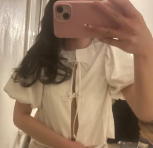 Vit blus med puffärm och knytning - Supersöt vit blus med puffiga korta ärmar och två knytband framtill som ger en trendig look. Blusen har en öppen detalj framtill och en mjuk, lätt struktur.