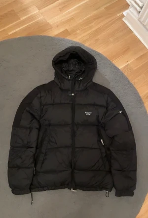 Svart pufferjacka från Alessandro Zavetti - Säljer en svart pufferjacka från Alessandro med huva och dragkedja framtill. Jackan har en clean look med diskret logga på bröstet och justerbara snören nedtill. Perfekt för kalla dagar när du vill hålla stilen enkel och snygg.