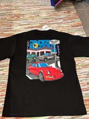 Svart Gallery Dept t-shirt med biltryck - Svart t-shirt från Gallery Dept. med färgstark grafisk print på ryggen som visar en röd sportbil, SUV och pickup framför en bensinstation med palmer och solnedgång. Framtill finns tryckt logga och adress i gult och vitt. Rund hals och korta ärmar. Materialet är mjuk bomull.