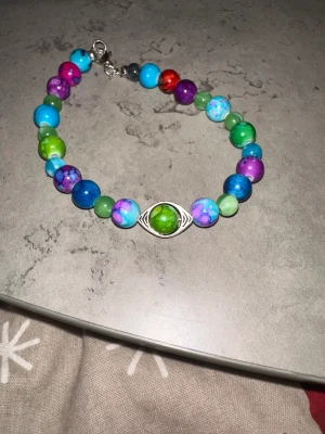 Hemma gjorda armband❣️gjord av kärlek trygghet och skydd -  Namn ”green.bluo.pink/Purple” artikelnummer ”2O11” 18 cm, Unikt armband med runda pärlor i blått, grönt, lila, rosa och rött. I mitten sitter en silverfärgad berlock med ett grönt öga. Pärlorna har marmorerad finish och armbandet har metallspänne. Perfekt för dig som gillar färg och coola detaljer. 