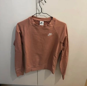 Rosa sweatshirt från Nike - Säljer en snygg rosa sweatshirt från Nike med klassisk logga broderad i vitt på bröstet. Tröjan har rund hals, långärmade raglanärmar och är gjord i mjuk bomullsmix. Perfekt för chill eller streetwear-stil.