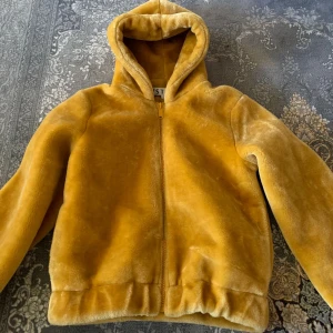 Gul teddyjacka med huva - Mysig gul teddyjacka med huva och dragkedja framtill. Jackan har en fluffig känsla och är perfekt för dig som vill ha en varm och trendig look. Passar dig som gillar streetstyle och vill sticka ut med färg.