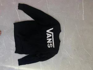 Svart sweatshirt från Vans - Svart sweatshirt från Vans i storlek medium med stort vitt Vans-tryck på bröstet. Tröjan har rund halsringning, långa ärmar och ribbade muddar. Tillverkad i mjukt material, perfekt för en avslappnad streetstil.