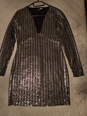 Glittrig silverklänning från H&M Divided - Snygg kort glittrig klänning från H&M Divided i storlek M. Klänningen har lång ärm, djup v-ringning och är täckt av silverfärgade paljetter i ett randigt mönster. Perfekt för dig som vill sticka ut med en festlig look.