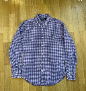 3-4skjortor   - Tre skjortor från Ralph Lauren i klassisk stil. En lila-rutig med vit knappstängning, en blå-rutig med tunna linjer och en blå-vit randig. Alla har lång ärm, button-down krage och broderad logga på bröstet. Skjortorna är gjorda i mjuk bomull och har normal passform.