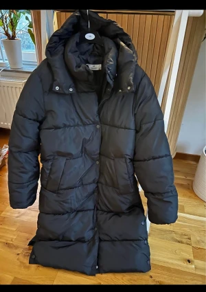 Svart lång pufferjacka med huva storlek 158 - Säljer en svart lång pufferjacka från H&M med stor huva och rejäla fickor. Jackan har quiltad design, tryckknappar och dragkedja framtill. Perfekt för kalla dagar och har en riktigt chill oversized vibe.