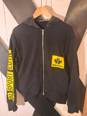  DSQUARED2 hoodie  - Svart hoodie från DSQUARED2 med gul DSQUARED2-logga och lönnlöv på bröstet samt gul text '04 SPORT EDTN' längs ena ärmen. Dragkedja framtill och huva i glansigt material. Snygg streetstil med coola detaljer och ribbade muddar. Skick 10-10. Nypris 3000kr Mitt pris 399kr Storlek 16Y Motsvarar Xs. Vid frågor eller funderingar hör av er 📩