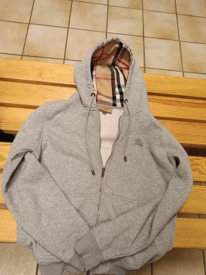 Grå Burberry hoodie med rutig foder - Grå hoodie från Burberry med klassiskt rutigt foder i huvan och insidan. Tröjan har dragkedja, snörning vid huvan och broderad Burberry-logga på bröstet. Materialet är mjuk bomull och polyester, perfekt för chilliga dagar. Snygg och tidlös design med långärmade ärmar.