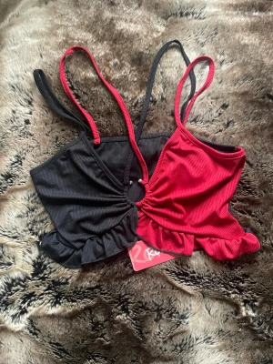 Svart och röd ribbad linnetopp med volang - Cool tvåfärgad linnetopp från Ragged Priest i svart och rött. Toppen har smala axelband, ribbad struktur och en rund metallring i mitten som delar färgerna. Volangkant nedtill ger en extra touch. Perfekt för dig som gillar statement-plagg!