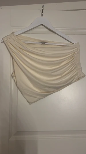 Beige oneshoulder topp med drapering - Säljer en beige oneshoulder topp med snygg drapering och rynkade detaljer längs ena sidan. Toppen är axelbandslös på ena axeln och har en cool asymmetrisk form som ger en trendig vibe. Perfekt för dig som gillar stilrena och moderna plagg.