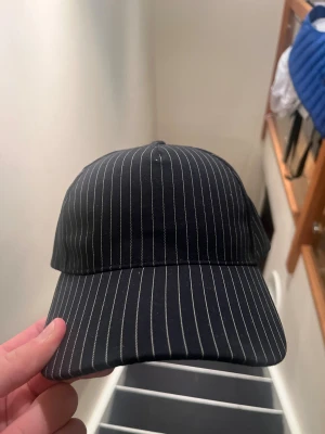 Svart keps Jean Paul Gaultier exklusiv - Snygg svart keps från Jean Paul Gaultier Parfums med tunna vita vertikala ränder. Klassisk böjd skärm och justerbar rem bak. Märkespatch baktill. Tillverkad i mjukt tyg, perfekt för dig som gillar street och designer.