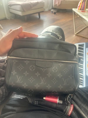 Louis Vuitton svart axelväska monogram - Snygg svart axelväska från Louis Vuitton med klassiskt monogrammönster i grått. Väskan har justerbar axelrem, dragkedja framtill och är tillverkad i tåligt skinn. Perfekt för dig som vill ha en stilren och ikonisk accessoar med plats för det viktigaste.