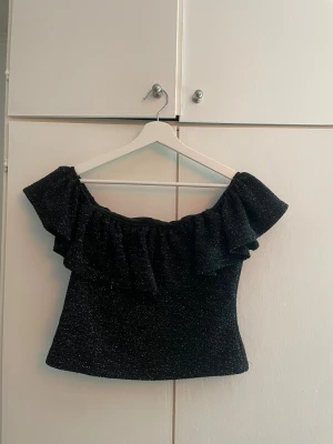Glittrig nyårstopp - Säljer en svart glittrig offshoulder topp från H&M i storlek M. Toppen har bred volang längs axlarna och är axelbandslös med en croppad passform. Materialet är mjukt och stretchigt med silverglitter i tyget som ger en festlig vibe. Passar mig me xs/s också 