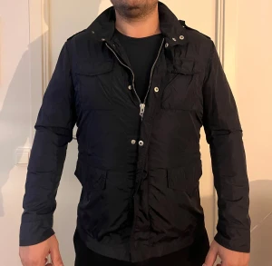 Snygg Gant jacka med fickor! - Snygg svart vindjacka med hög krage och flera praktiska fickor framtill. Jackan har dragkedja och tryckknappar, samt axelklaffar som ger en cool touch. Passformen är normal och materialet känns lätt och smidigt, perfekt för blåsiga dagar.