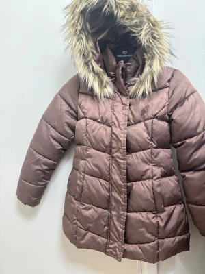 Brun pufferjacka med fuskpäls från H&M - Snygg brun pufferjacka från H&M med fluffig fuskpäls runt huvan. Jackan är quiltad och har långa ärmar, dragkedja och tryckknappar framtill. Perfekt för kalla dagar och har en lite längre passform som håller dig varm.