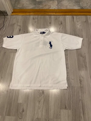 Vit Polo Ralph Lauren piké XL - Vit pikétröja från Polo Ralph Lauren i bomull, med klassisk krage och knappar framtill. Stor mörkblå polospelare broderad på bröstet och två polospelare på ryggen. Snygg detalj med siffra på ärmen och logga i kragen. Perfekt för en clean och sportig look.