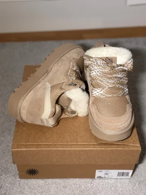 Uggs lowmel  - Snygga beige sneakers från UGG med grov sula, snörning och insida i mjuk fårull. Ovandel i mocka och textil, detaljerade mönstrade skosnören och UGG-logga på sidan. Perfekta för kyliga dagar när du vill vara både varm och trendig.