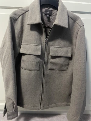 Beige overshirt jacka från H&M - Snygg grå overshirt jacka från H&M med klassisk krage och två stora bröstfickor med lock. Jackan har dragkedja framtill och är tillverkad i mjukt ullblandat material. Perfekt för lager-på-lager och har en clean, loose passform.