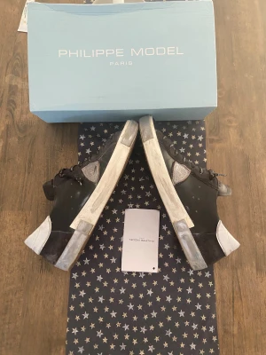 Svarta Philippe Model sneakers - Säljer ett par svarta Philippe Model sneakers med vita detaljer och logotyp på sidan. Skorna har snörning med svarta och vita skosnören med text, rund tå och platt sula. Materialet är skinn och syntet, vilket ger en cool och lyxig känsla. Perfekt för dig som gillar streetstyle.