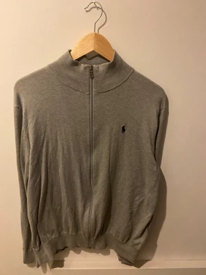 Grå zip-kofta från Polo Ralph Lauren - Snygg grå kofta från Polo Ralph Lauren i mjuk pima bomull. Tröjan har hel dragkedja framtill, ribbad krage och muddar samt den klassiska Polo-loggan broderad på bröstet. Perfekt för chill dagar och enkel att matcha med jeans eller chinos.