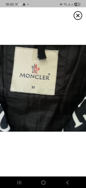 Svart jacka från Moncler i storlek M - Snygg svart jacka från Moncler i storlek M. Jackan har en klassisk look med Moncler-logga på insidan och är tillverkad i ett lätt, glansigt material. Perfekt för dig som vill ha en stilren och exklusiv jacka med streetkänsla.