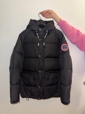 Svart pufferjacka från Tuxer M - Svart pufferjacka från Tuxer med huva och snörning, storlek M. Jackan har ett broderat märke på ärmen, stora fickor framtill och knappar längs öppningen. Perfekt för kalla dagar med sin tjocka och quiltade design. Snygg och funktionell look. Mycket fint skick, är sparsamt använd.