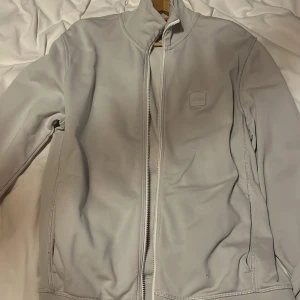 Ljusgrå vindjacka från Hugo boss - Full zip Hugo boss, Perfekt för dig som gillar minimalistisk look och sportig stil. Har ett litet hål i fram väldigt svårt att se och går att sy