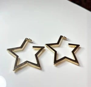 Stjärnformade örhängen  - Säljer ett par stora, stjärnformade örhängen i metall med guldig finish. De har en cool, kantig design och fästs med klassisk stift. Perfekta för dig som vill sticka ut och addera lite extra bling till din look.