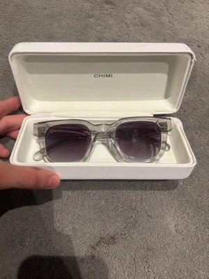 Chimi transparenta solglasögon - Snygga solglasögon från Chimi med transparenta, fyrkantiga bågar i plast och silvriga detaljer på skalmarna. Glasen är mörklila och har en cool gradient-effekt. Kommer med vit hård förvaringsbox. Designade i Stockholm.