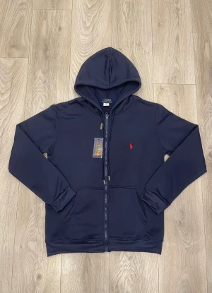 Marinblå hoodie från Polo Ralph Lauren - Snygg marinblå hoodie från Polo Ralph Lauren med dragkedja och huva. Tröjan har ett rött broderat Polo-logo på bröstet, praktiska fickor framtill och ribbade muddar. Tillverkad i mjuk bomullsmix som är skön att ha på sig. Perfekt för chill dagar.