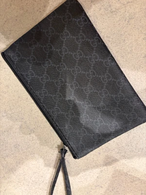 Svart kuvertväska f - Svart kuvertväska från  med klassiskt över hela ytan. Väskan har en stilren rektangulär form, dragkedja upptill och en handledsrem. Materialet är slitstark syntet med en lyxig känsla. Perfekt för att bära dina essentials med stil.