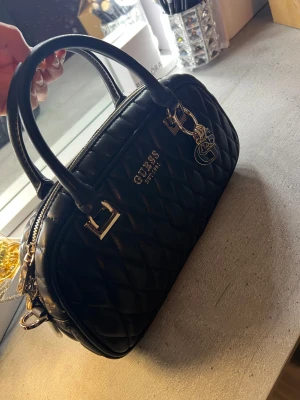 Svart quiltad handväska från Guess - Snygg svart handväska från Guess med quiltad design och guldfärgade detaljer. Väskan har både handtag och avtagbar axelrem, samt en dekorativ Guess-nyckelring. Insidan är ljus med logomönster och har en praktisk dragkedjeficka💕.