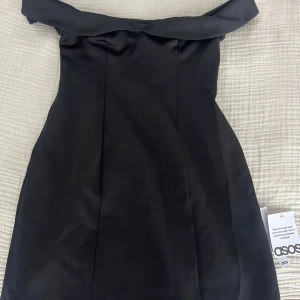 Svart offshoulder klänning från ASOS - Helt oanvänd supersnygg svart kortklänning från ASOS med offshoulder-design och v-ringad överdel. Klänningen har en stilren, figurnära passform upptill och lätt utsvängd kjol. Perfekt för dig som vill sticka ut med en elegant och modern look. Har även samma klänning i storlek 36. Ordinariepris 399kr.