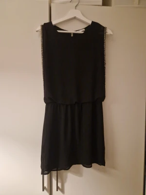 Svart kort klänning med pärldetaljer - Svart kort klänning från C i storlek 38. Klänningen har ärmlös design och är dekorerad med snygga pärldetaljer längs axlarna. Den har en avslappnad passform med markerad midja och är tillverkad i ett lätt, flowy material.