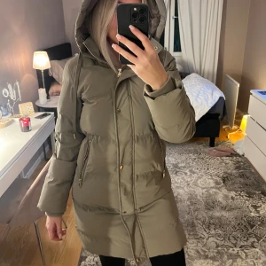 Vinterjacka - Säljer en lång beige pufferjacka med huva och snörning. Knappt använd. Jackan har dragkedja och knappar framtill, samt två fickor med guldfärgade dragkedjor. Perfekt för kalla dagar och har en snygg, enkel design som passar till det mesta.