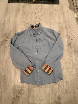 Blå Burberry Brit skjorta XS - Snygg ljusblå skjorta från Burberry Brit i bomull, med klassisk krage och knappar framtill. Insidan av kragen och manschetterna har det ikoniska rutiga Burberry-mönstret i beige, svart och rött. Perfekt för dig som gillar stilrena detaljer och designerplagg.